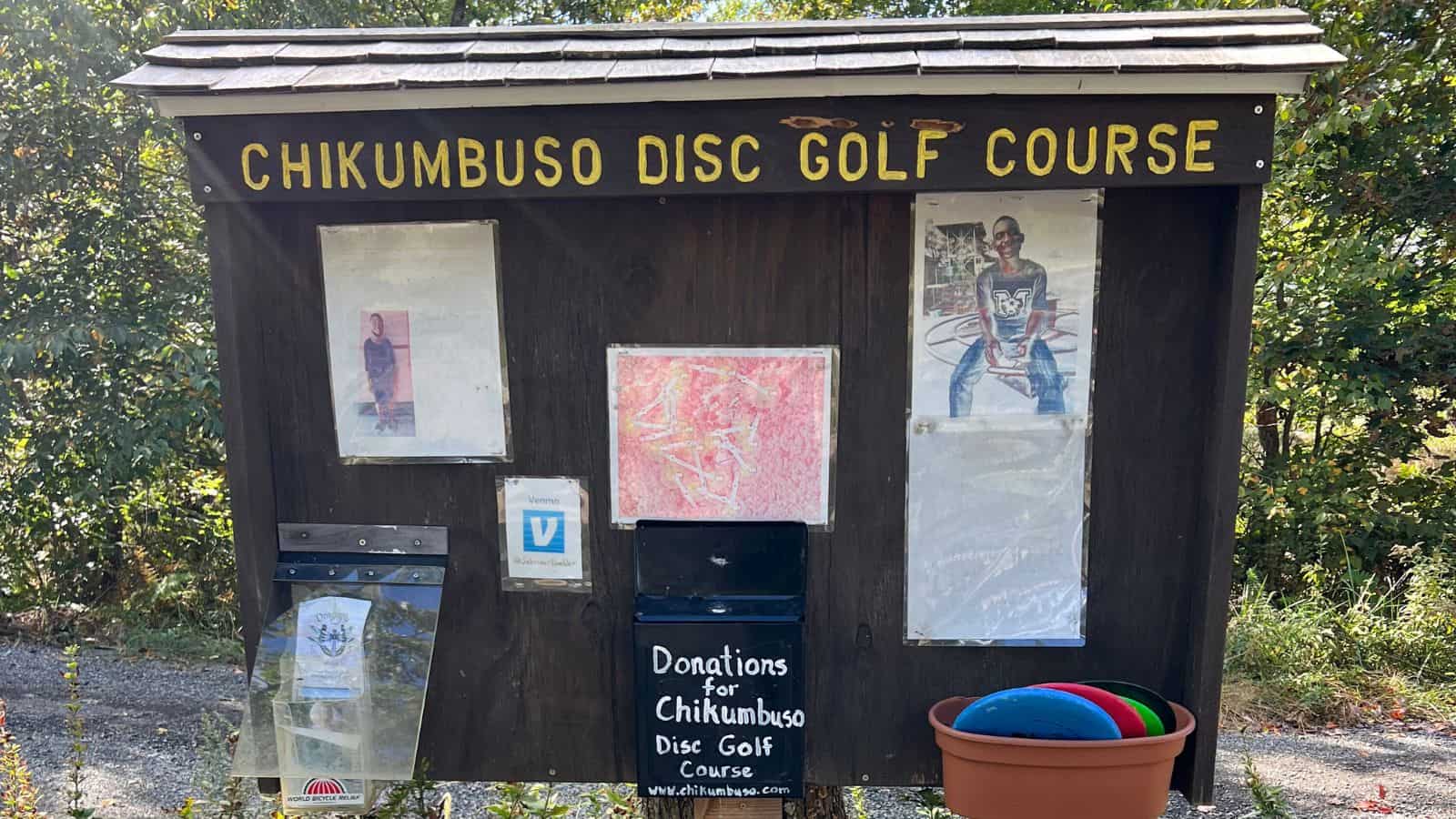 Wooden signboard for Chikumbuso Disc Golf Course with donation box, course information, and a bin containing colorful discs, set outdoors with trees in the background.
