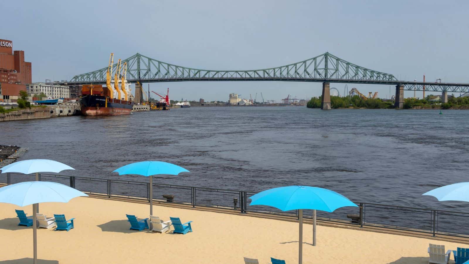 A sandy urban beach with blue umbrellas and chairs faces a wide river, with a large steel bridge and docked cargo ships in the background.