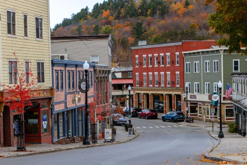 A quiet small town street in Saranac Lake, lined with colorful buildings, autumn trees, parked cars, and a backdrop of forested hills ablaze with fall foliage.