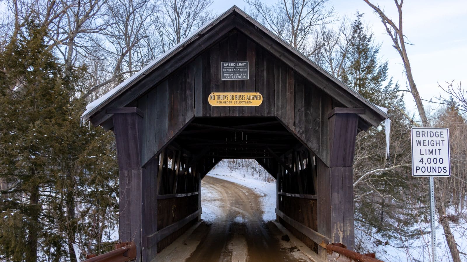 A wooden covered bridge over a dirt road—one of the haunted places around New England—with signage indicating a speed and 4,000-pound weight limit; trees and snow create an eerie backdrop.
