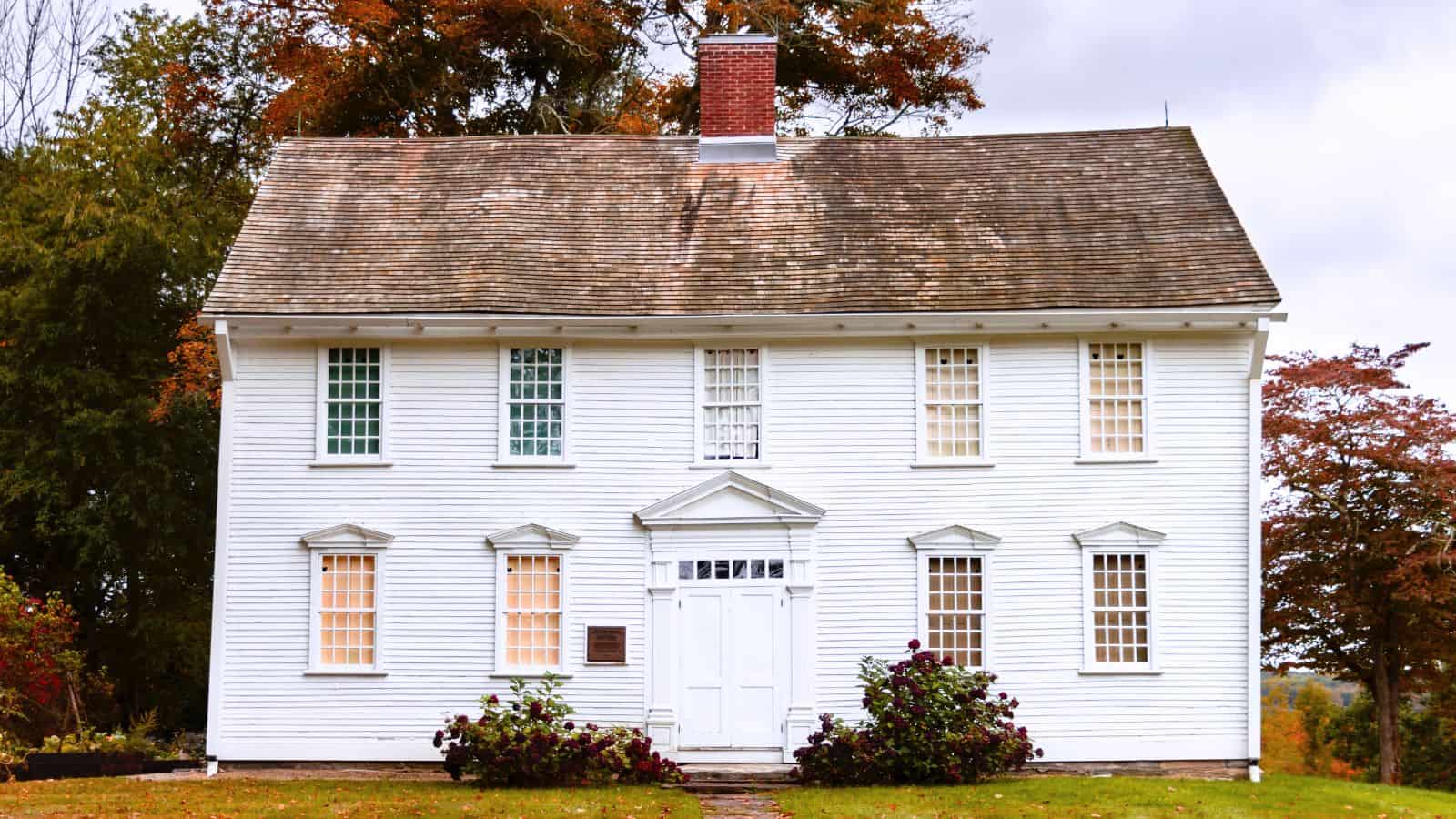 A two-story white colonial-style house with a steep roof, six front windows, a centered door, and autumn trees in the background.