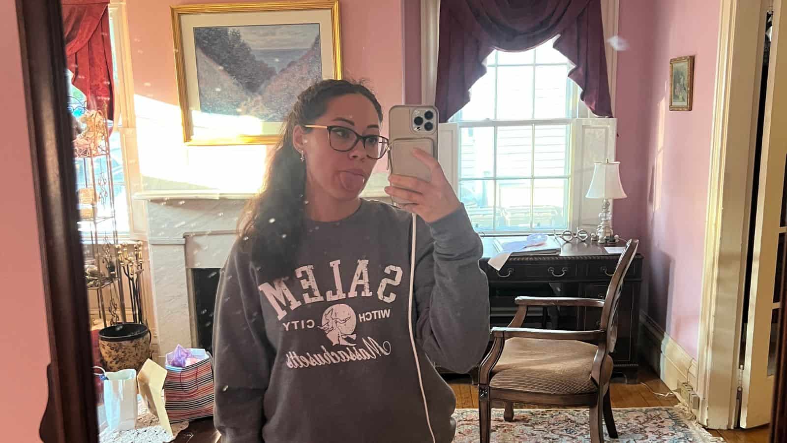 Woman taking a mirror selfie in a pink room, wearing glasses and a "Salem Witch City" sweatshirt, sticking out her tongue. A desk, chair, and window are in the background.