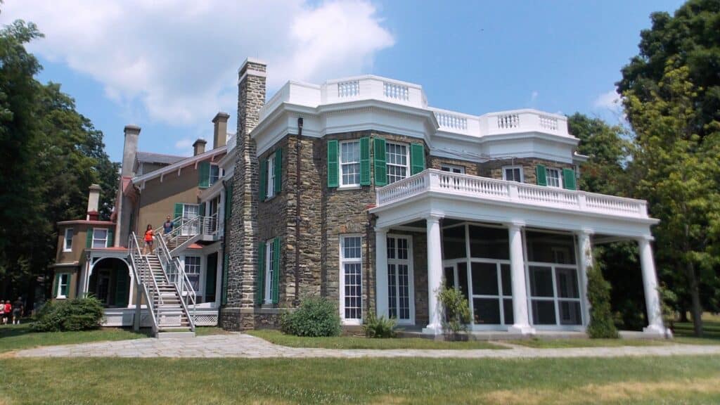 A large historic mansion with stone walls, green shutters, and a white columned porch, surrounded by trees and a lawn under a partly cloudy sky.