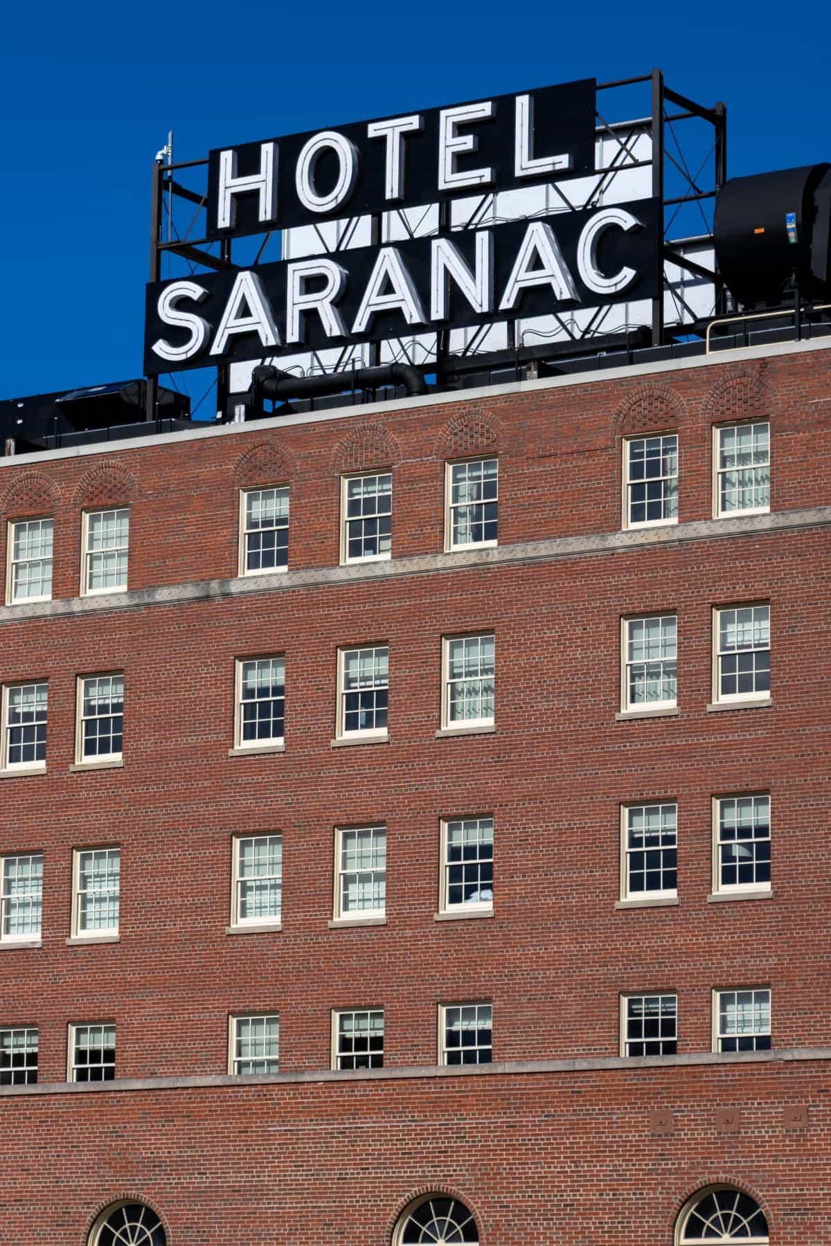 A brick building with multiple windows and a large rooftop sign that reads "HOTEL SARANAC" stands proudly against a clear blue sky in the heart of Saranac Lake.