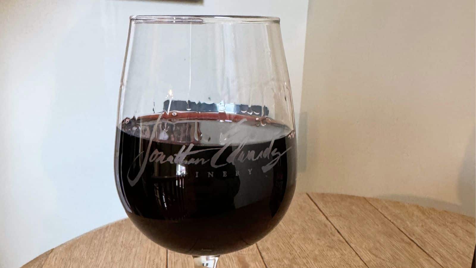 A glass of red wine sits on a wooden table, with "Jonathan Charles Winery" etched on the glass.