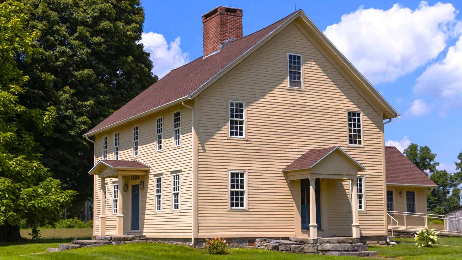 A two-story beige colonial-style house with a red brick chimney, multiple grid windows, and two entryways, set on a lawn with trees and a blue sky background.