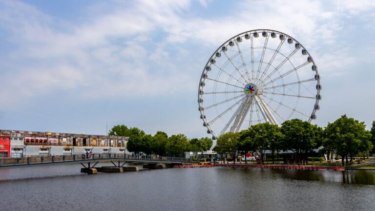A large Ferris wheel stands near a waterfront, with trees, a footbridge, and a few people visible under a partly cloudy sky.