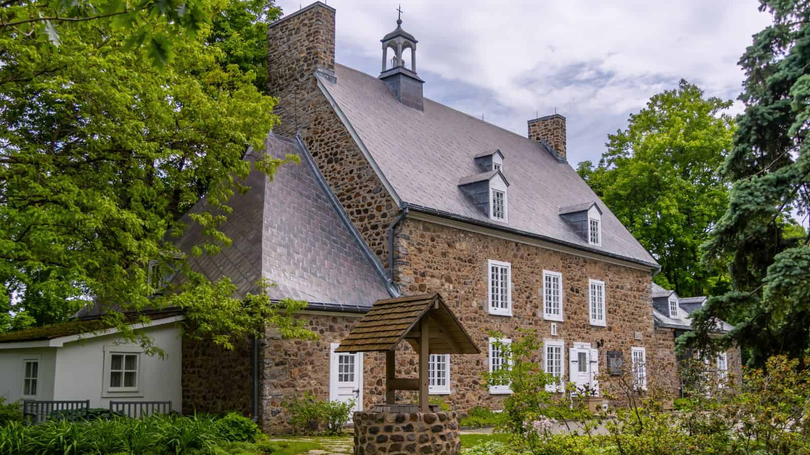 A large stone house with a steep roof, multiple chimneys, white-framed windows, and a wooden well in the front yard, surrounded by trees and greenery.