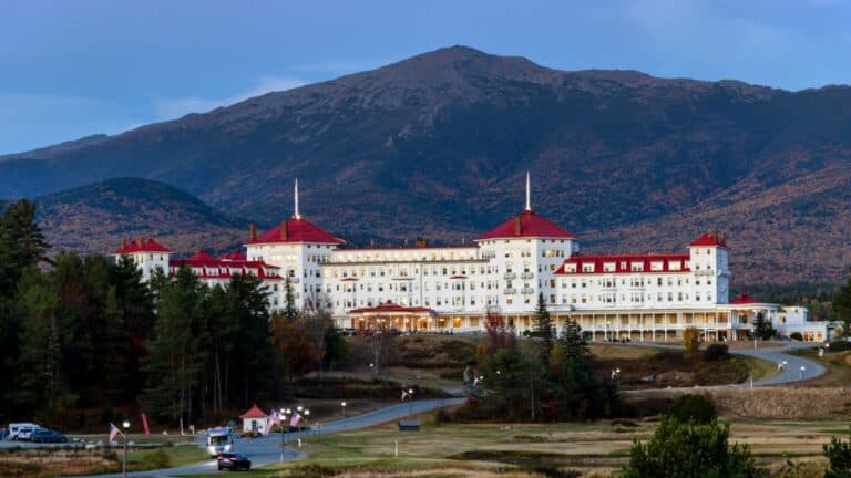 Large white hotel with red roofs set against a backdrop of forested hills and a tall mountain under a clear sky.