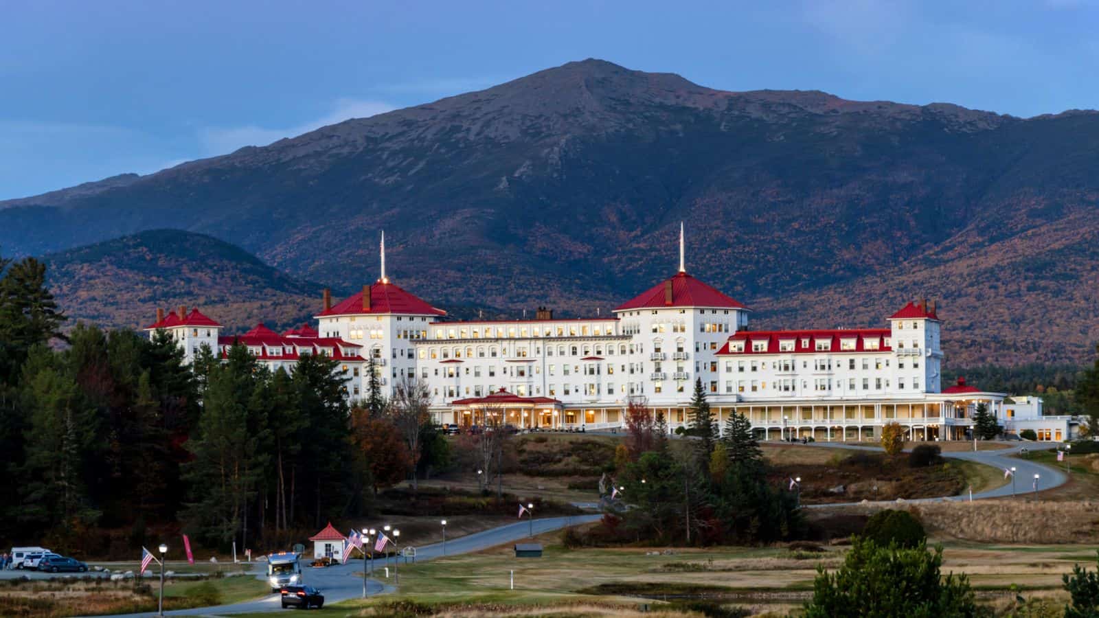 Large white hotel with red roofs set against a backdrop of forested hills and a tall mountain under a clear sky.