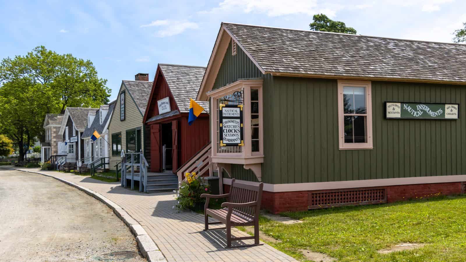 A row of historic wooden buildings with signs and small flags, along a paved walkway with benches and grass on a sunny day.