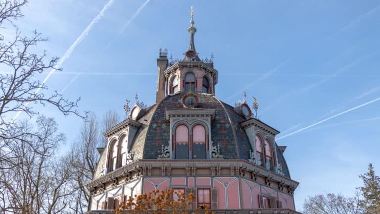 A multistory octagonal building with ornate detailing, pink accents, and round windows, set against a clear blue sky with visible airplane contrails.