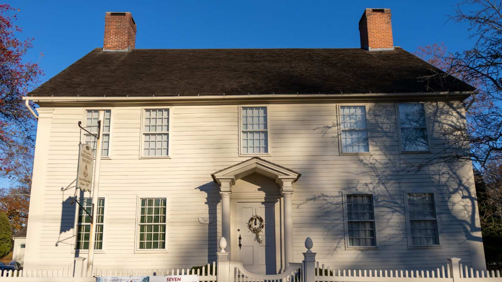 A two-story white colonial-style house with symmetrical windows, two chimneys, and a central front door, surrounded by a white picket fence.