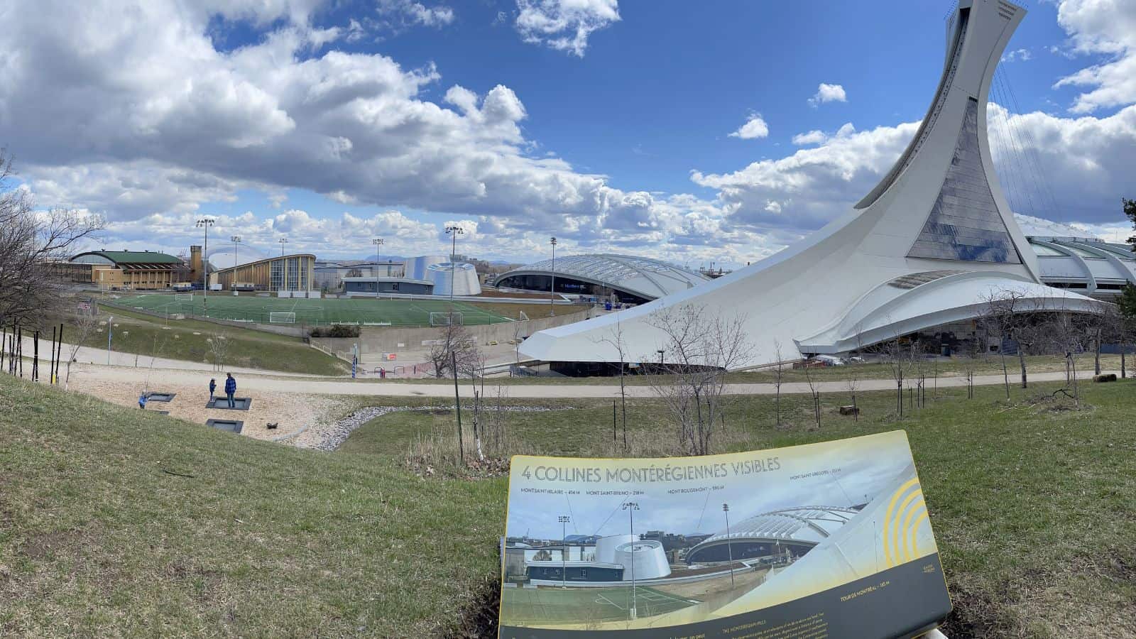 Wide-angle view of Montreal’s Olympic Stadium exterior with a bilingual informational sign about Montérégiennes hills in the foreground, under a partly cloudy sky.