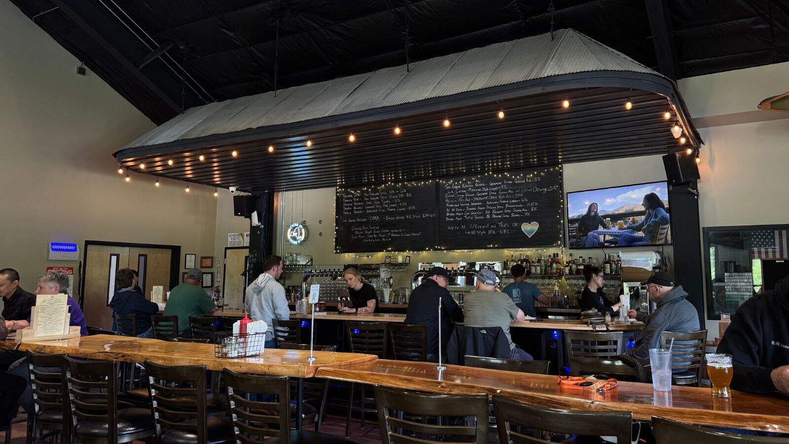 People sit at a bar with a wooden counter, overhead string lights, a large menu board, and a TV on the wall showing two people on a talk show.