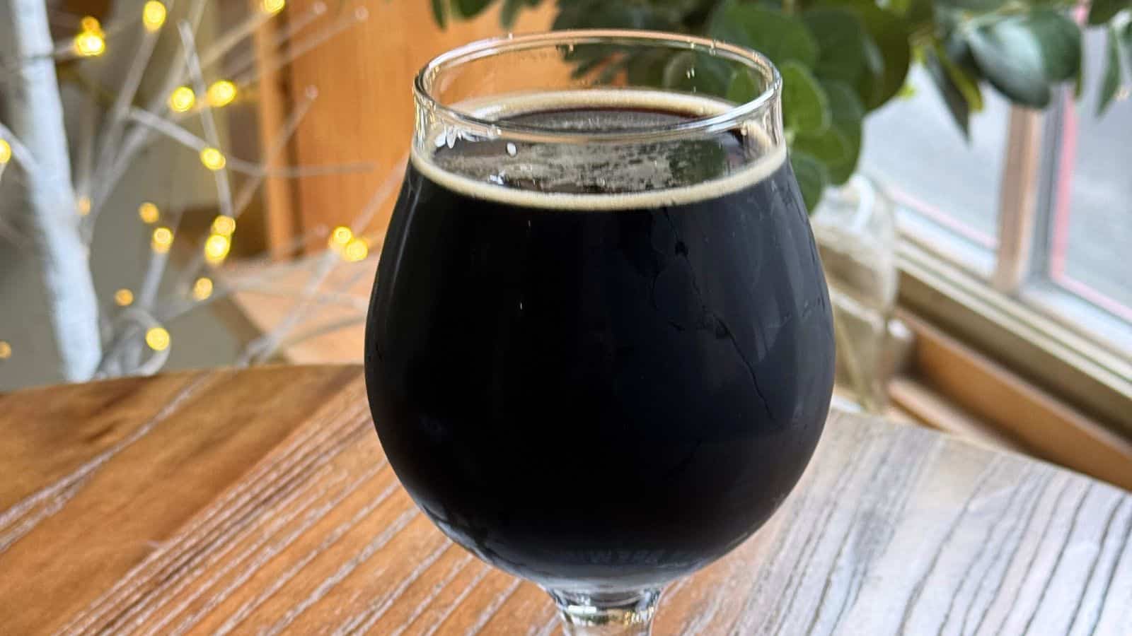 A glass of dark beer on a wooden table near a window, with plants and warm string lights in the background.