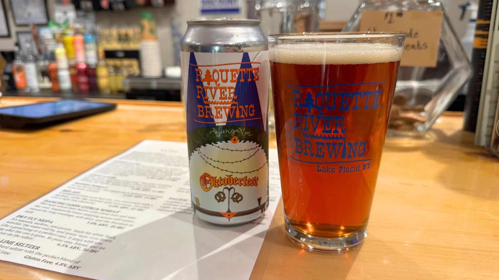 A can and a pint glass of Raquette River Brewing Oktoberfest beer sit on a bar counter next to a printed menu.