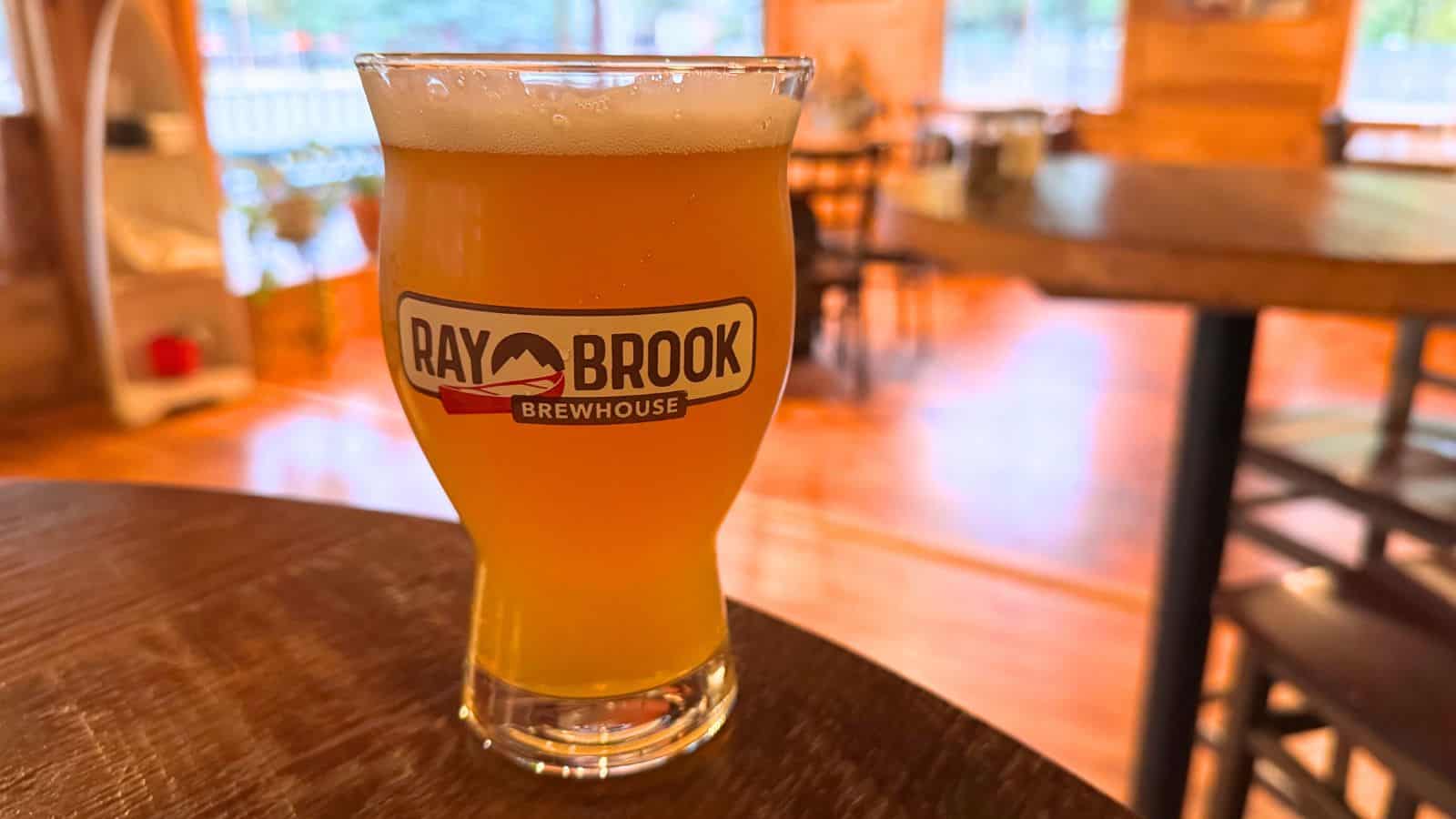 A pint glass of beer with a foamy head, labeled "Ray Brook Brewhouse," sits on a table in a rustic indoor setting.