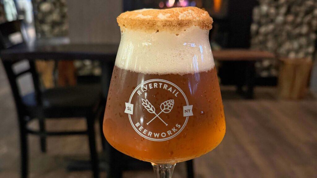 A glass of amber beer from Rivertrail Beerworks with a foamy head and a sugared rim sits on a table in a dimly lit room.