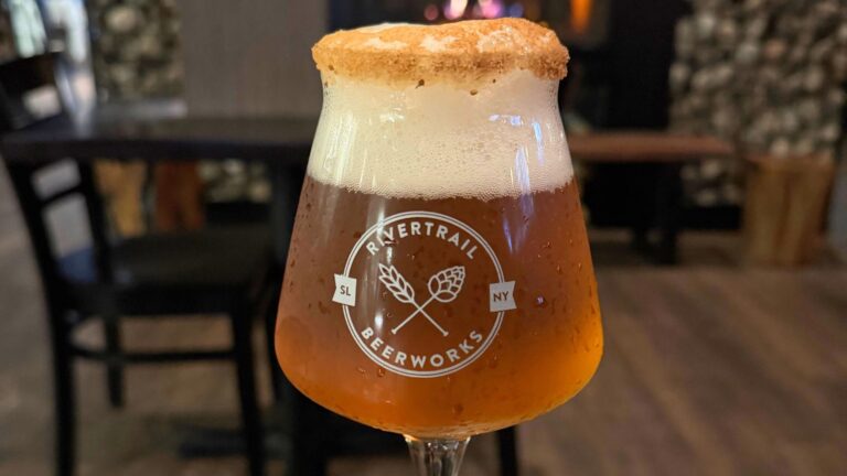 A glass of amber beer from Rivertrail Beerworks with a foamy head and a sugared rim sits on a table in a dimly lit room.