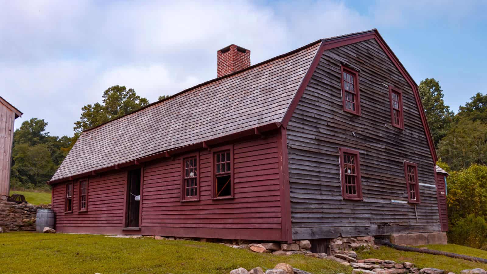 A long, red wooden house with a gabled roof, multiple windows, and a brick chimney, set on a grassy area with trees in the background.