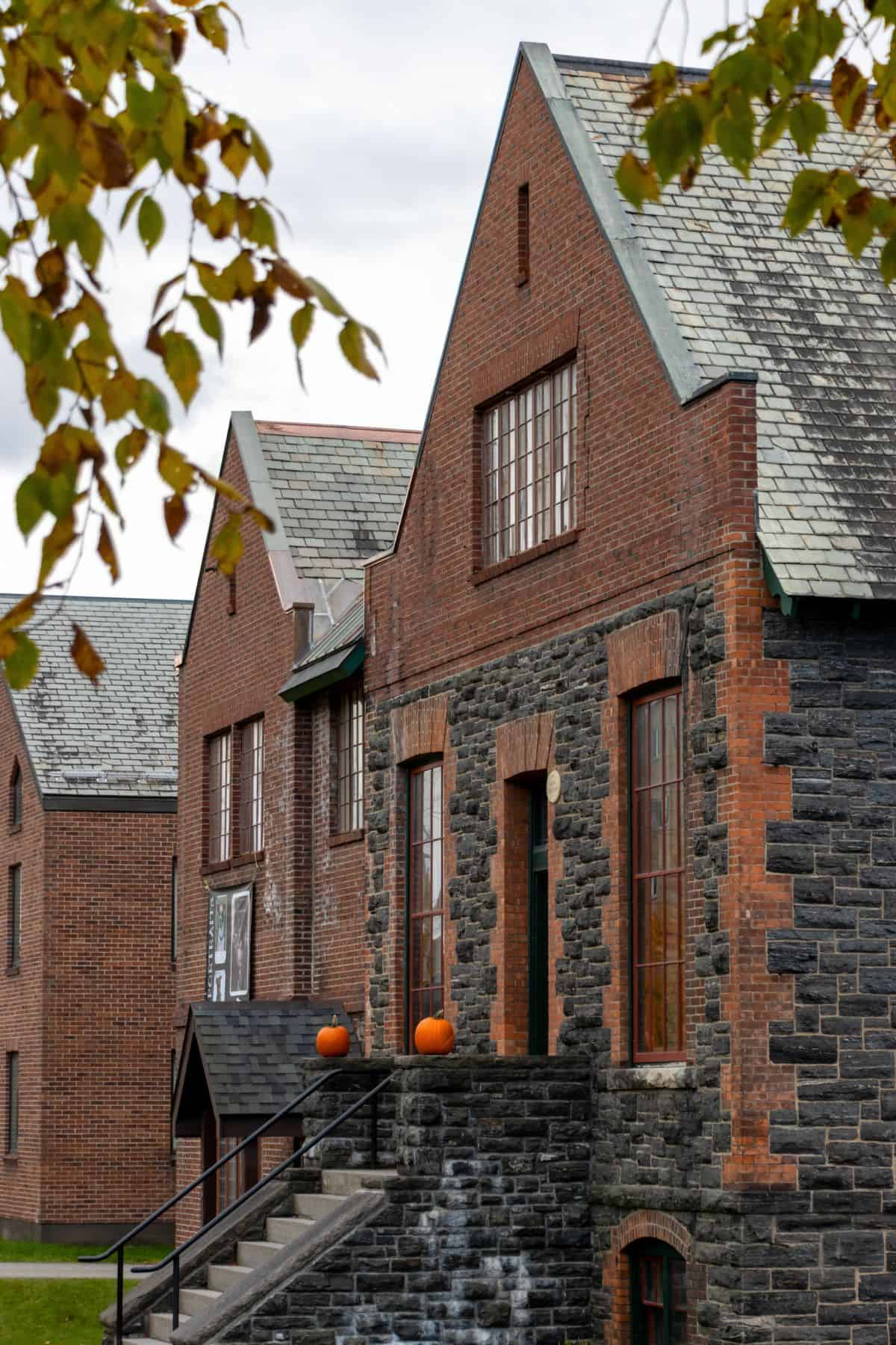 Three brick and stone buildings with pitched roofs in Saranac Lake; two pumpkins sit on the stairs leading to the entrance. Tree branches with green leaves frame the image.