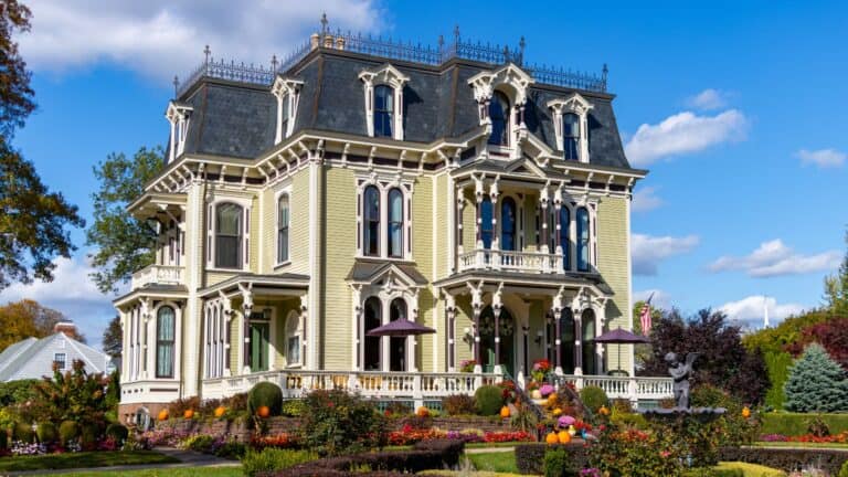 A large, ornate Victorian-style house with a mansard roof, surrounded by colorful gardens and landscaping under a blue sky.