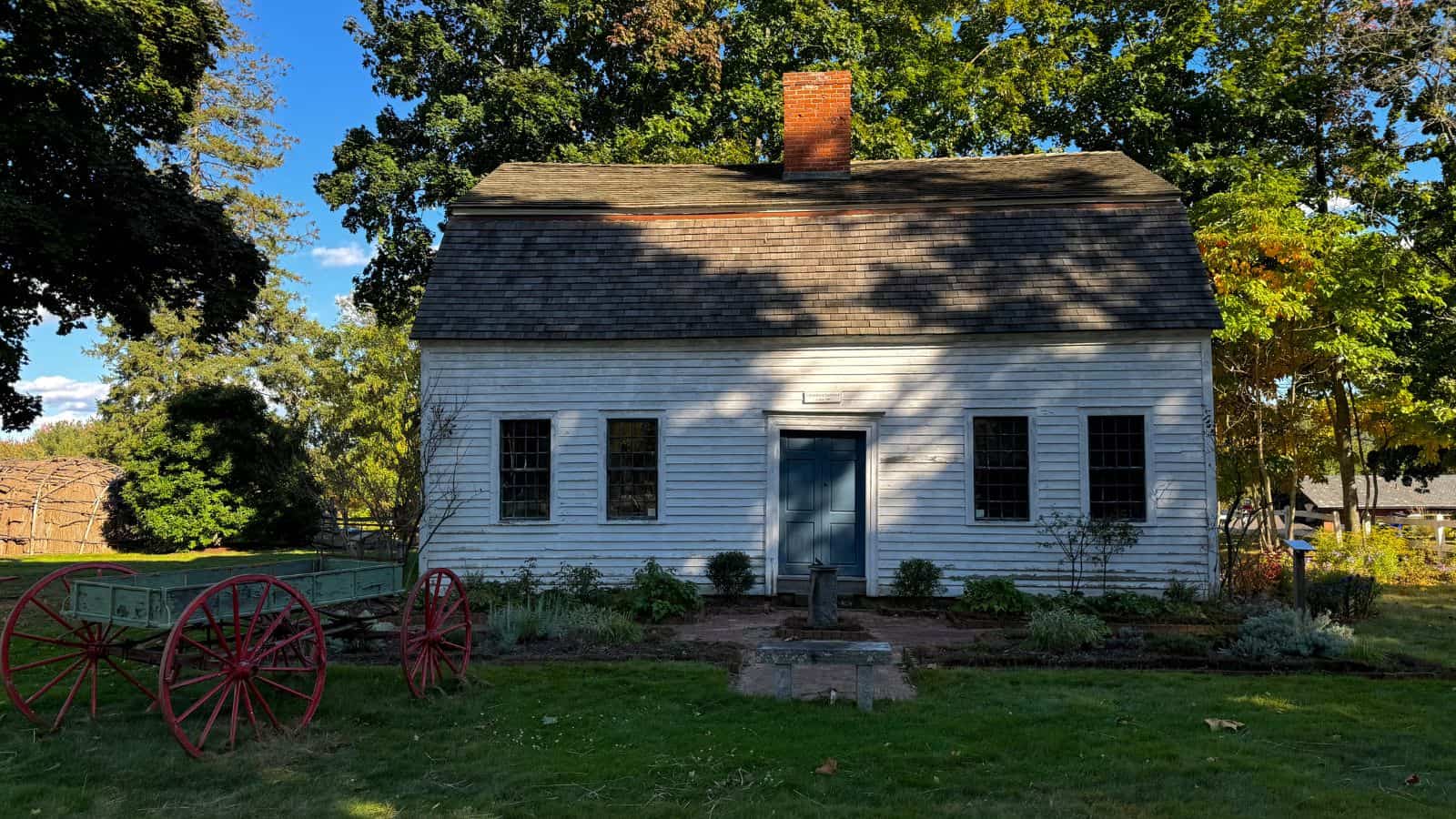 A small, white wooden house with a central brick chimney, six windows, and a blue door stands in front of a red-wheeled wooden wagon amid trees and greenery—evoking the charm of historic Connecticut towns.