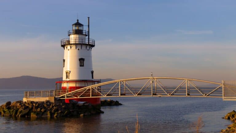 A white and red lighthouse stands on rocks by the water, connected to land by a metal bridge, with hills visible in the background under a clear sky.