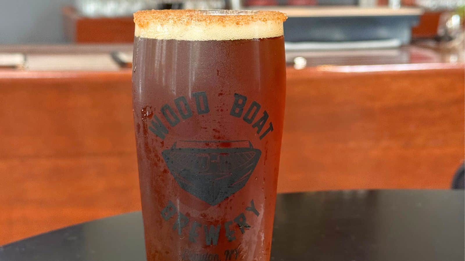A pint glass filled with dark beer, rimmed with what appears to be a spice mix, displaying the "Wood Boat Brewery" logo, sits on a dark table.