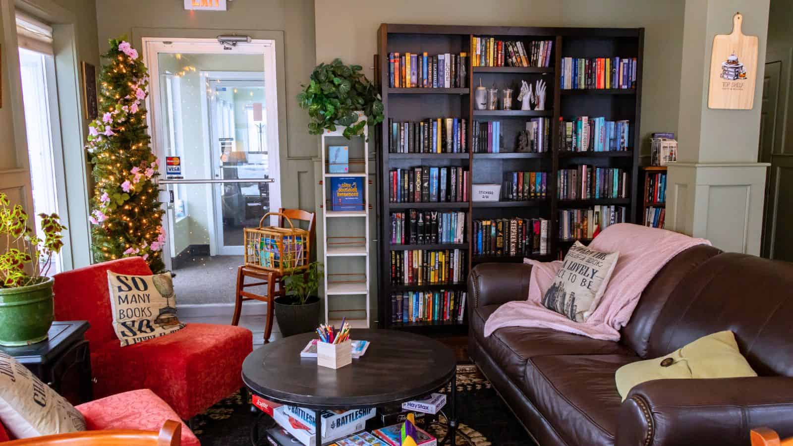 Cozy reading nook with a sofa, armchair, round table with games, bookshelves filled with books, and a small decorated tree near a glass door—perfect for relaxing after exploring things to do in North Stonington, Connecticut.