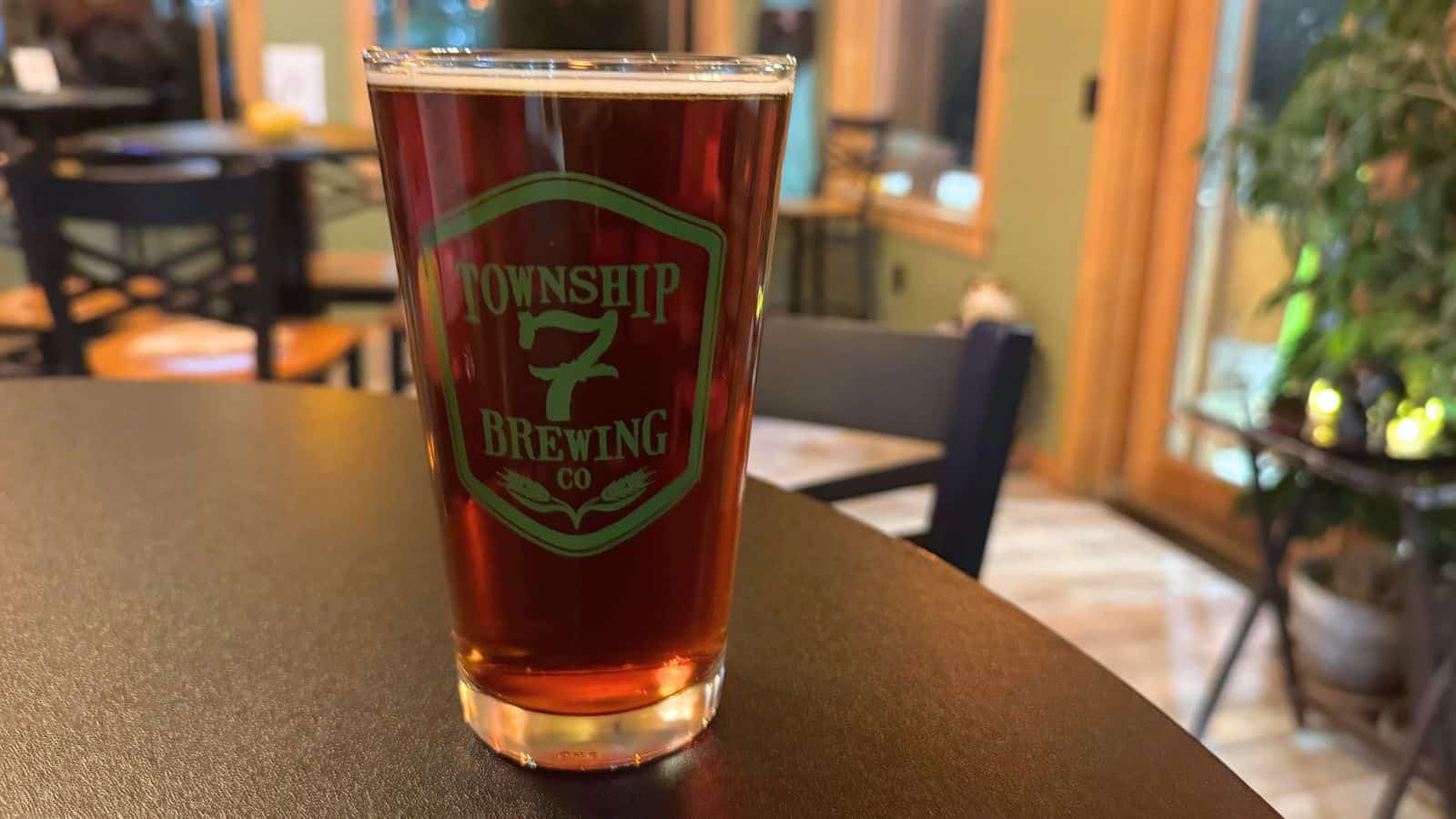 A pint glass filled with dark amber beer on a round table, featuring the Township 7 Brewing Co. logo, with chairs and plants in the background.