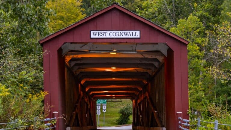 A red covered bridge with the sign "West Cornwall" above the entrance, surrounded by green trees and lit by interior lights—an iconic sight in one of the charming historic Connecticut towns.