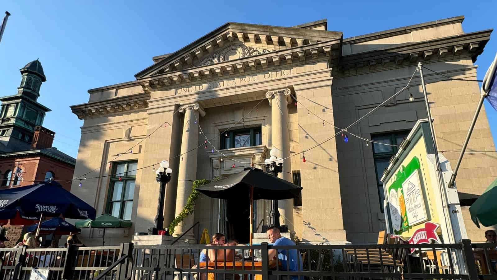 People sit at outdoor tables in front of a historic building labeled "United States Post Office" on a sunny day, with string lights and umbrellas—capturing the charm often found in historic Connecticut towns.