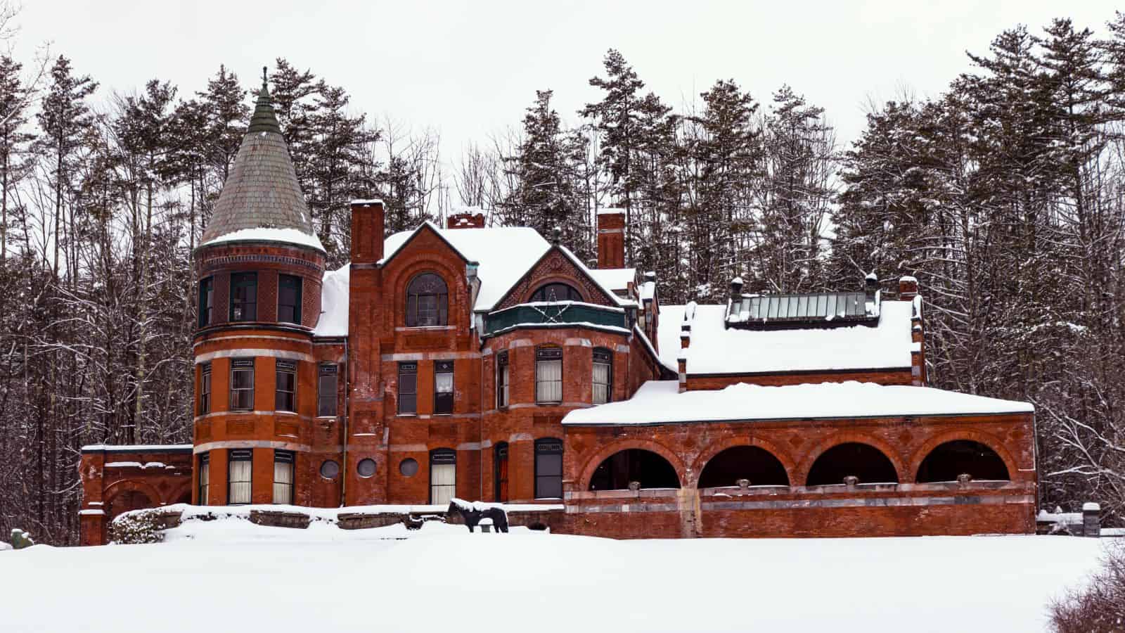 Large red-brick Victorian mansion with a round turret, arched porch, and snow-covered roof, situated in front of a forest of tall pine trees.