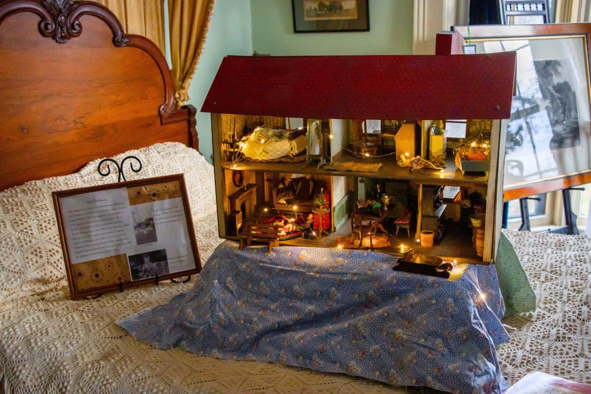 A detailed dollhouse is displayed on a bed covered with a patterned cloth, next to framed documents and photographs in a bedroom setting.