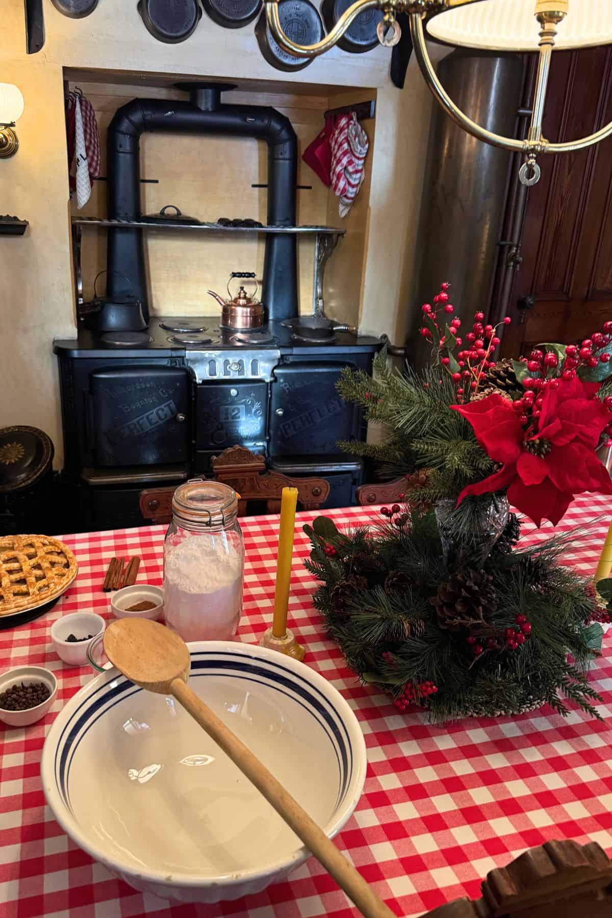 A vintage kitchen scene with a wood-burning stove, a checkered tablecloth, a bowl with a wooden spoon, baking ingredients, pies, and a festive holiday centerpiece.