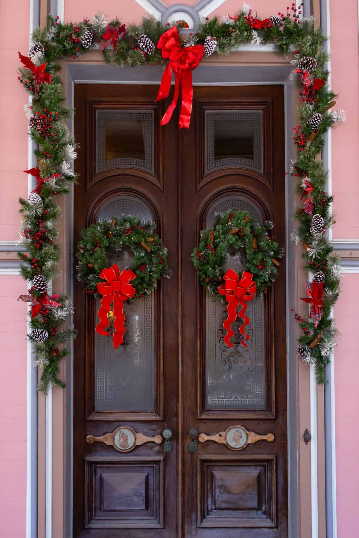 A wooden double door is decorated with two wreaths and garland featuring red bows, pine cones, and greenery for the Christmas holiday.