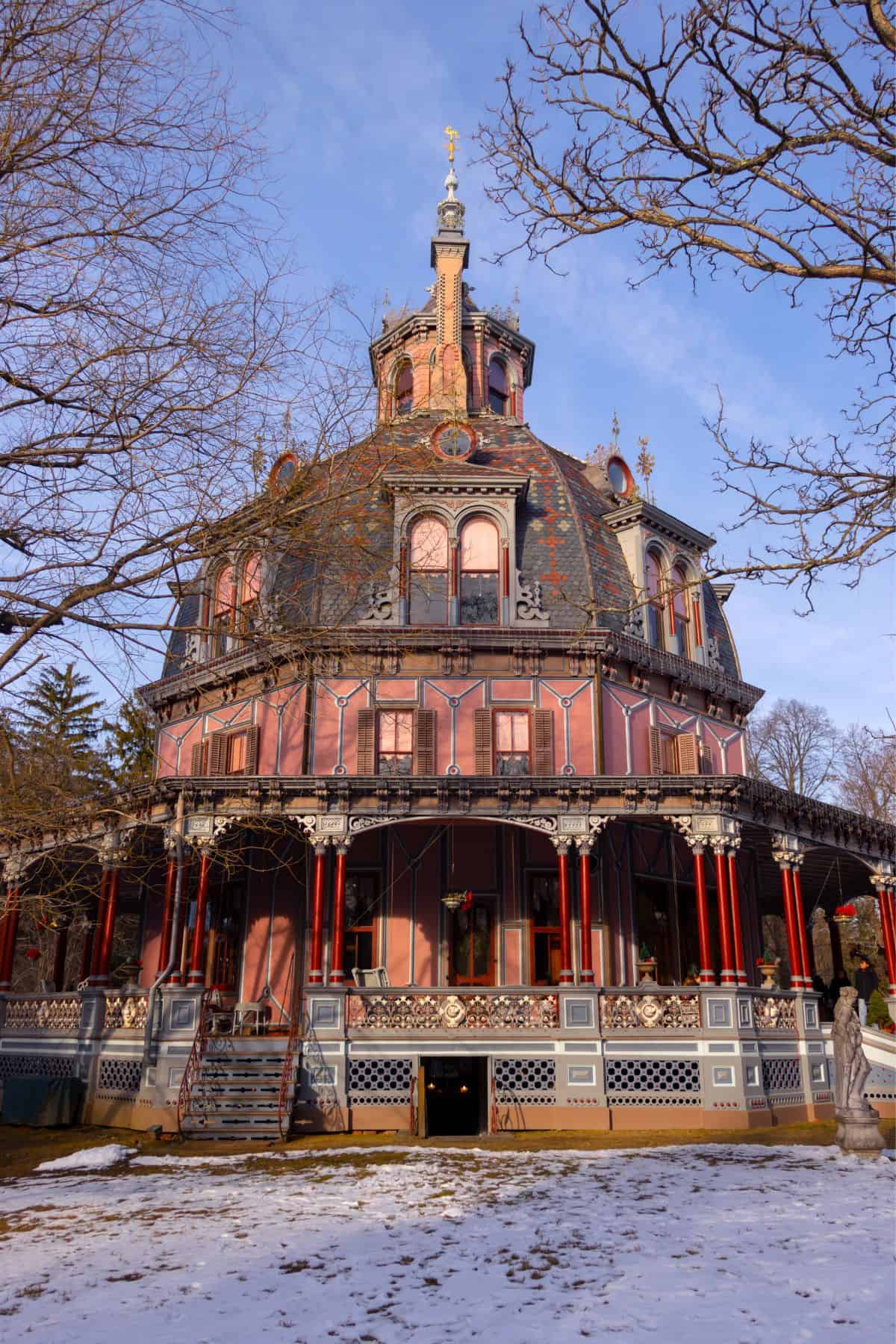 A large, ornate Victorian-style house with multiple levels, pink exterior walls, red columns, and intricate architectural details, surrounded by bare trees and some snow on the ground.