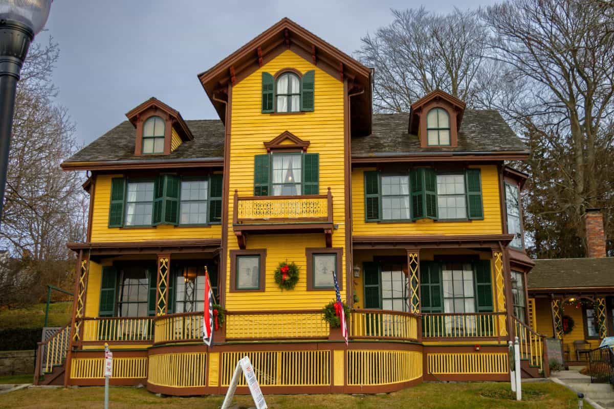 A large yellow two-story house with green shutters, a central balcony, and a decorated wreath above the entrance, surrounded by bare trees.