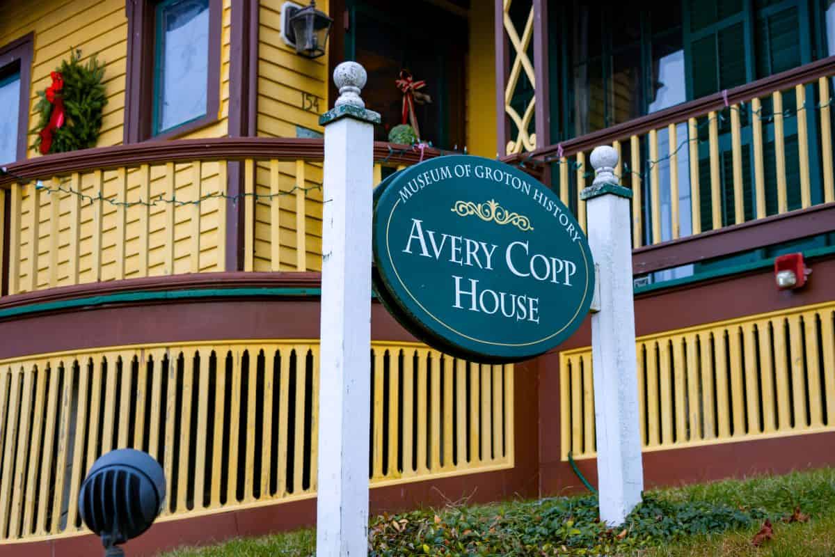 A green sign reading "Avery Copp House, Museum of Groton History" stands in front of a yellow house with a decorated porch.