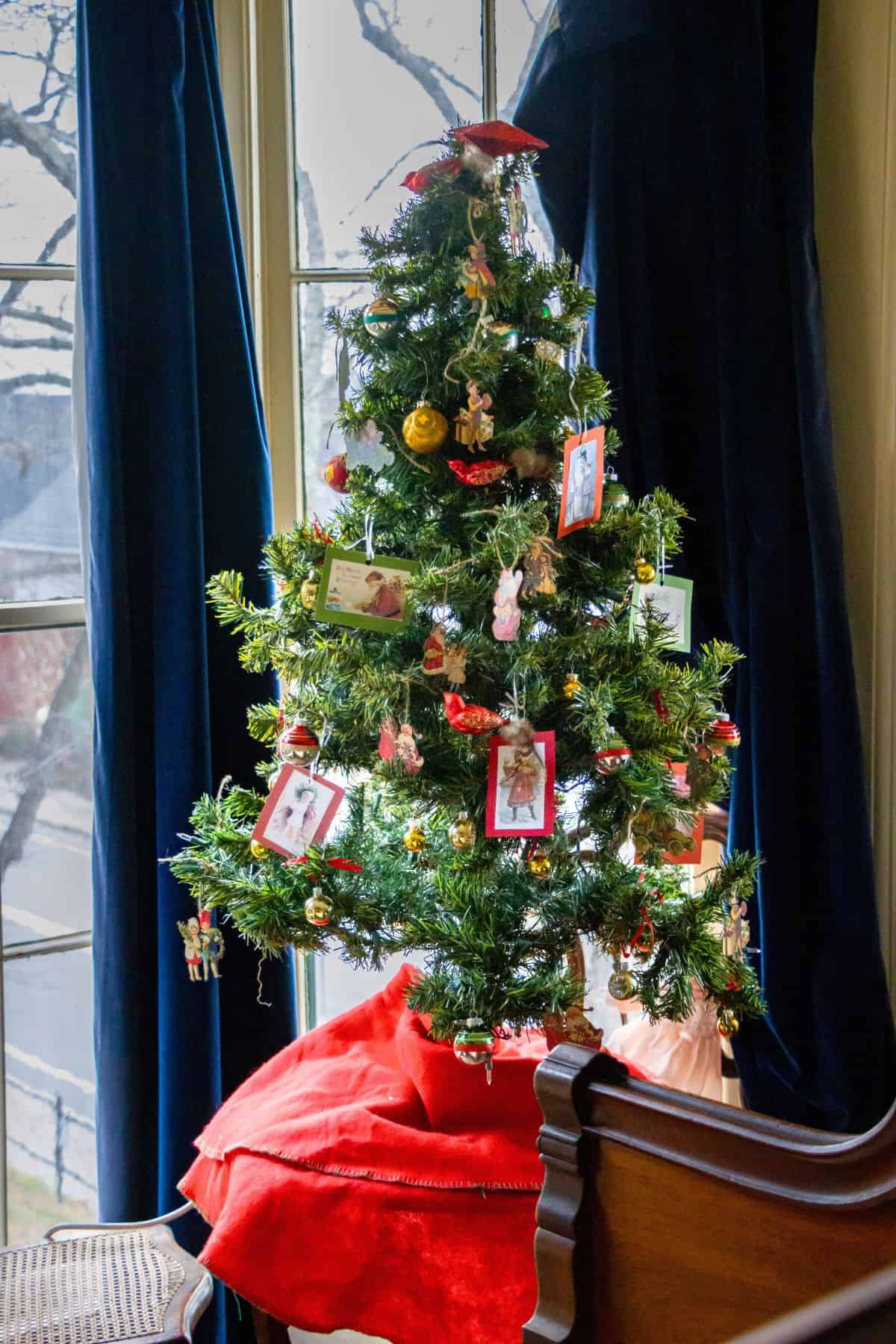 Small Christmas tree decorated with framed photos, ornaments, and ribbons, standing by a window with blue curtains and a red tree skirt at the base.