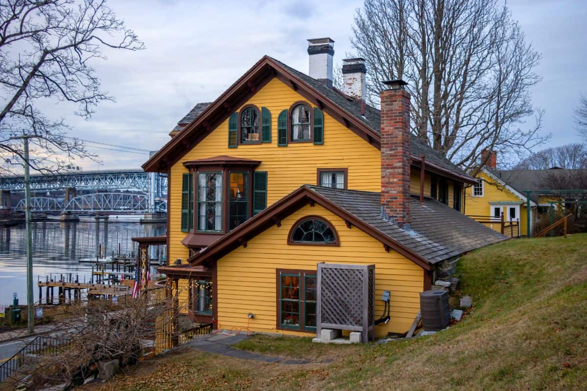 A yellow wooden house with green shutters sits on a sloped lawn next to a river, with a bridge visible in the background and trees without leaves surrounding the area.