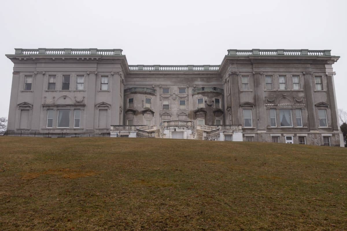 Large, gray, neoclassical-style mansion with stone facade and symmetrical wings, viewed from a grassy hill under an overcast sky.
