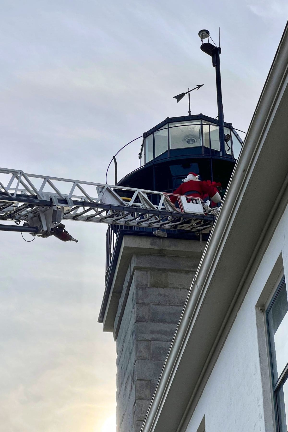 A person dressed as Santa Claus is being assisted by a fire truck ladder near the top of a lighthouse on a cloudy day.