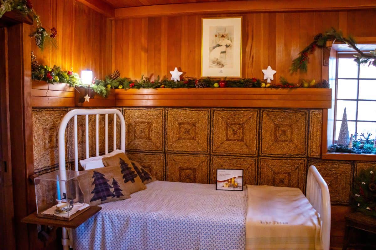A small bedroom with a single metal bed, pine and star decorations, wood-paneled walls, a window, and framed art above the bed.