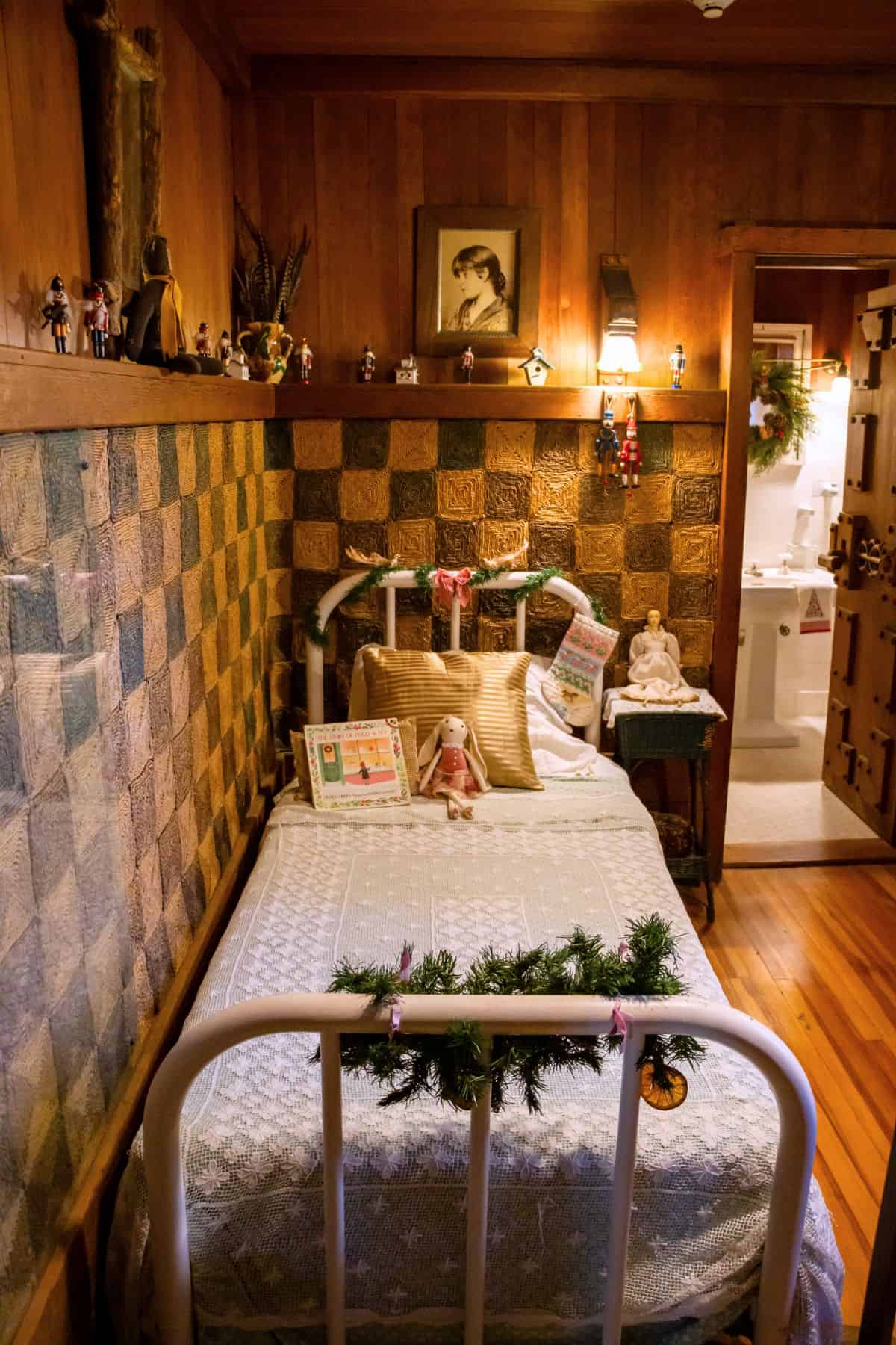 A small bedroom with a single bed, vintage decor, dolls, and holiday decorations; a bathroom is visible through an open door in the background.