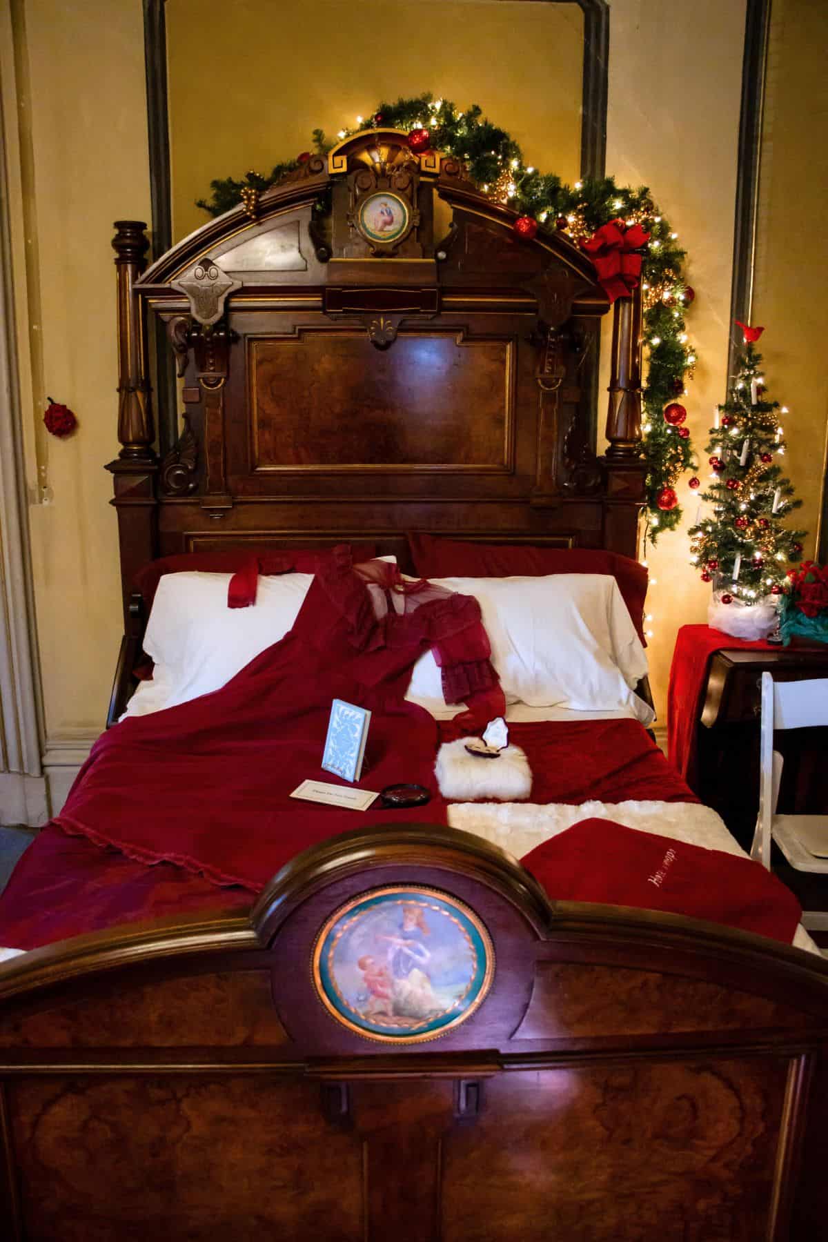 A vintage wooden bed with a red Victorian dress, a book, a stocking, and festive Christmas decorations, including garlands and a small decorated tree.