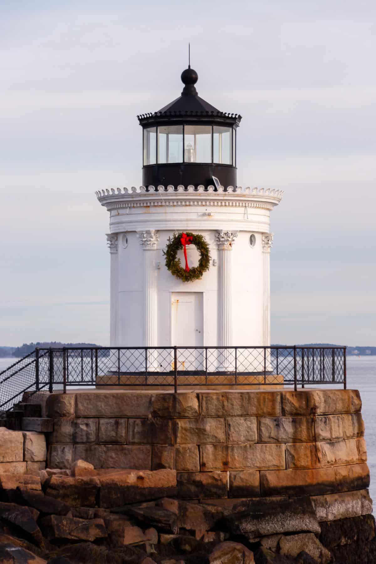 A white lighthouse with a black top stands on a stone foundation by the water, decorated with a green holiday wreath and red bow on its door.