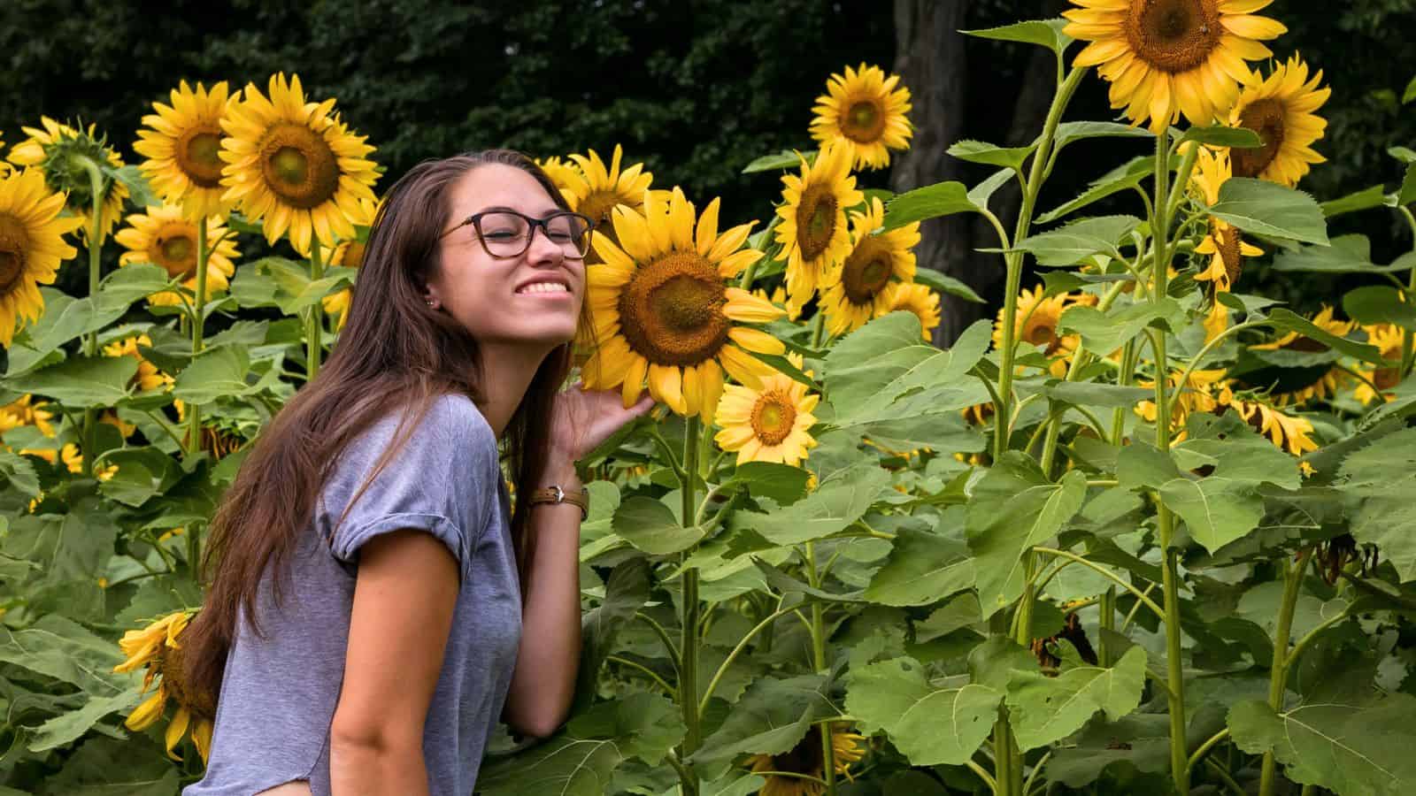 A woman with long brown hair and glasses smiles while standing among tall sunflowers in a garden.